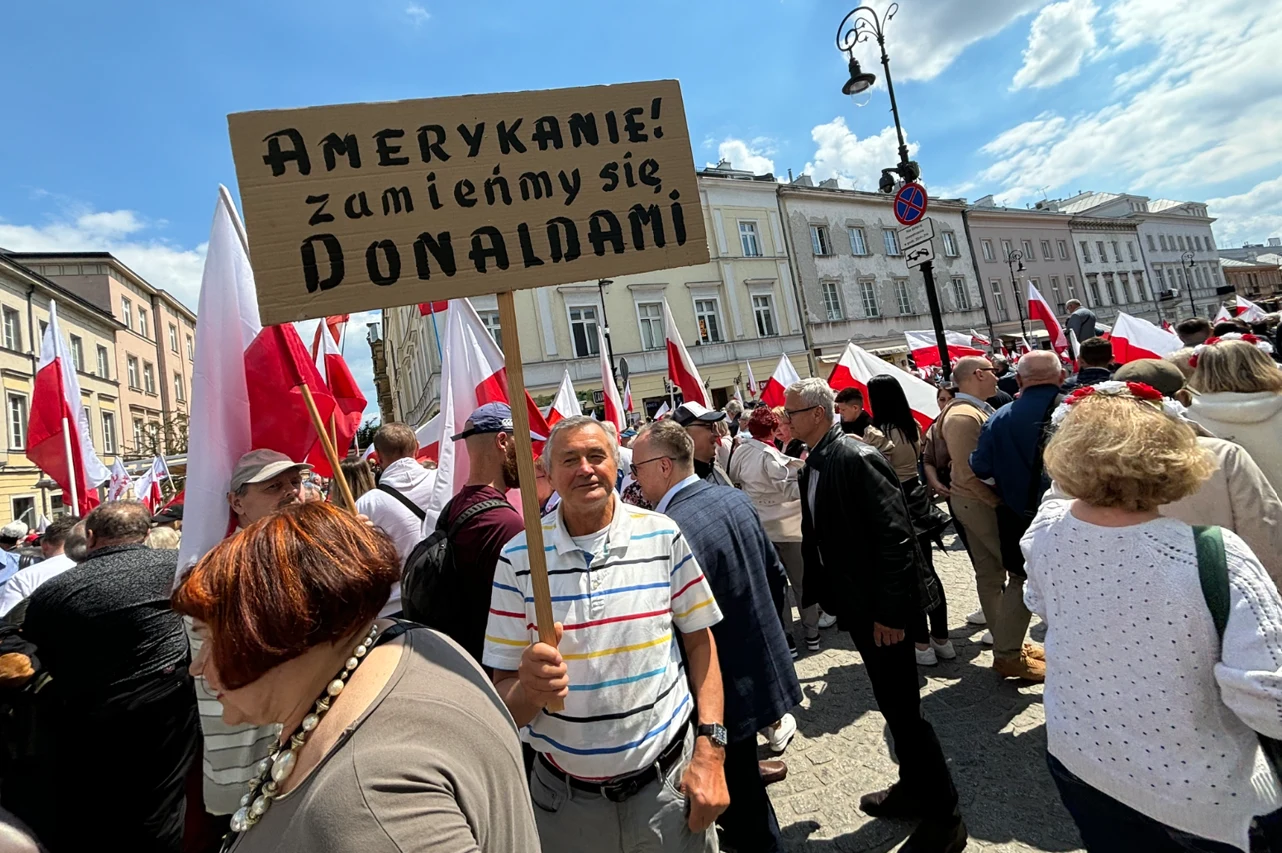 Uczestnicy manifestacji z flagami Polski na tle zabudowy miejskiej, mężczyzna na pierwszym planie trzyma tablicę z napisem 'AMERYKANIE! zamieńmy się DONALDAMI', wokół zgromadzenie ludzi obserwujących i biorących udział w wydarzeniu