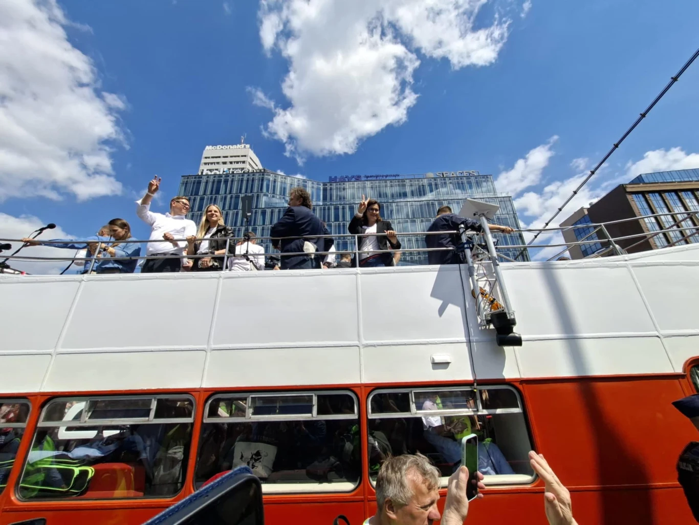 Ludzie stojący i siedzący na górnym pokładzie czerwonego, piętrowego autobusu w mieście pod błękitnym niebem z białymi chmurami, w tle nowoczesne przeszklone wieżowce.