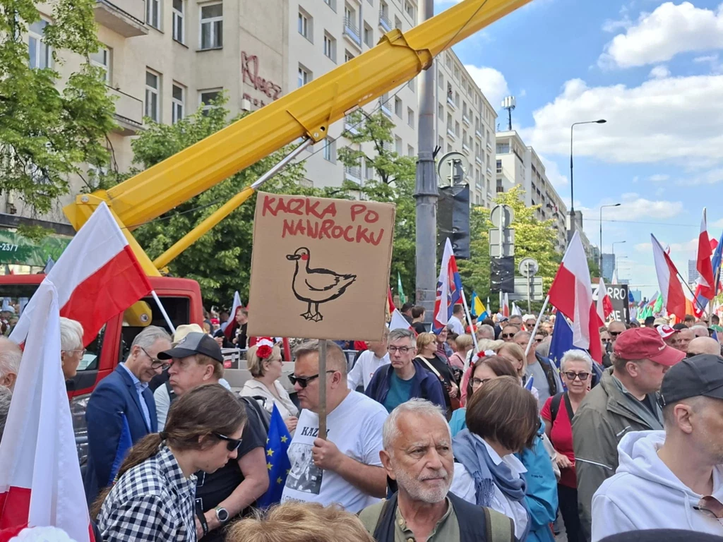 Nie zabrakło banerów uderzających w Karola Nawrockiego Grupa ludzi maszerująca ulicą podczas demonstracji, trzymających różne transparenty i flagi, w tym biało-czerwone flagi Polski, na pierwszym planie kartonowy plakat z rysunkiem kaczki i napisem 'Kaczka po Naukocku', w tle budynki mieszkalne i żółty dźwig.