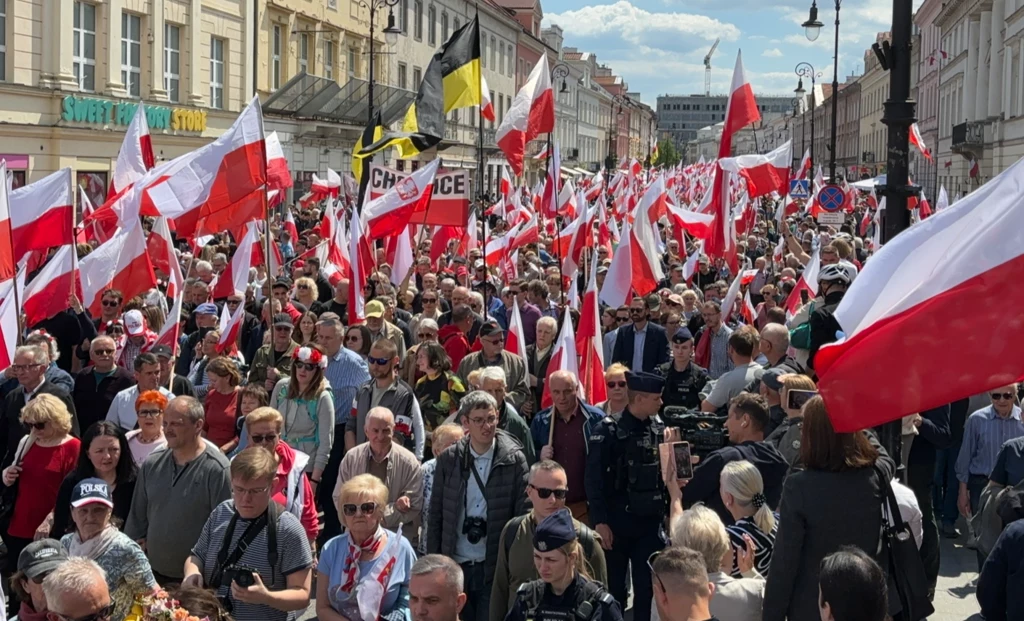 Nowy Świat szczelnie wypełniony uczestnikami Marszu za Polską Tłum ludzi maszeruje ulicą, większość uczestników trzyma polskie flagi, nad głowami góruje morze biało-czerwonych barw, po bokach stoją policjanci, a w tle widoczne są kamienice i niebo z lekkim zachmurzeniem.