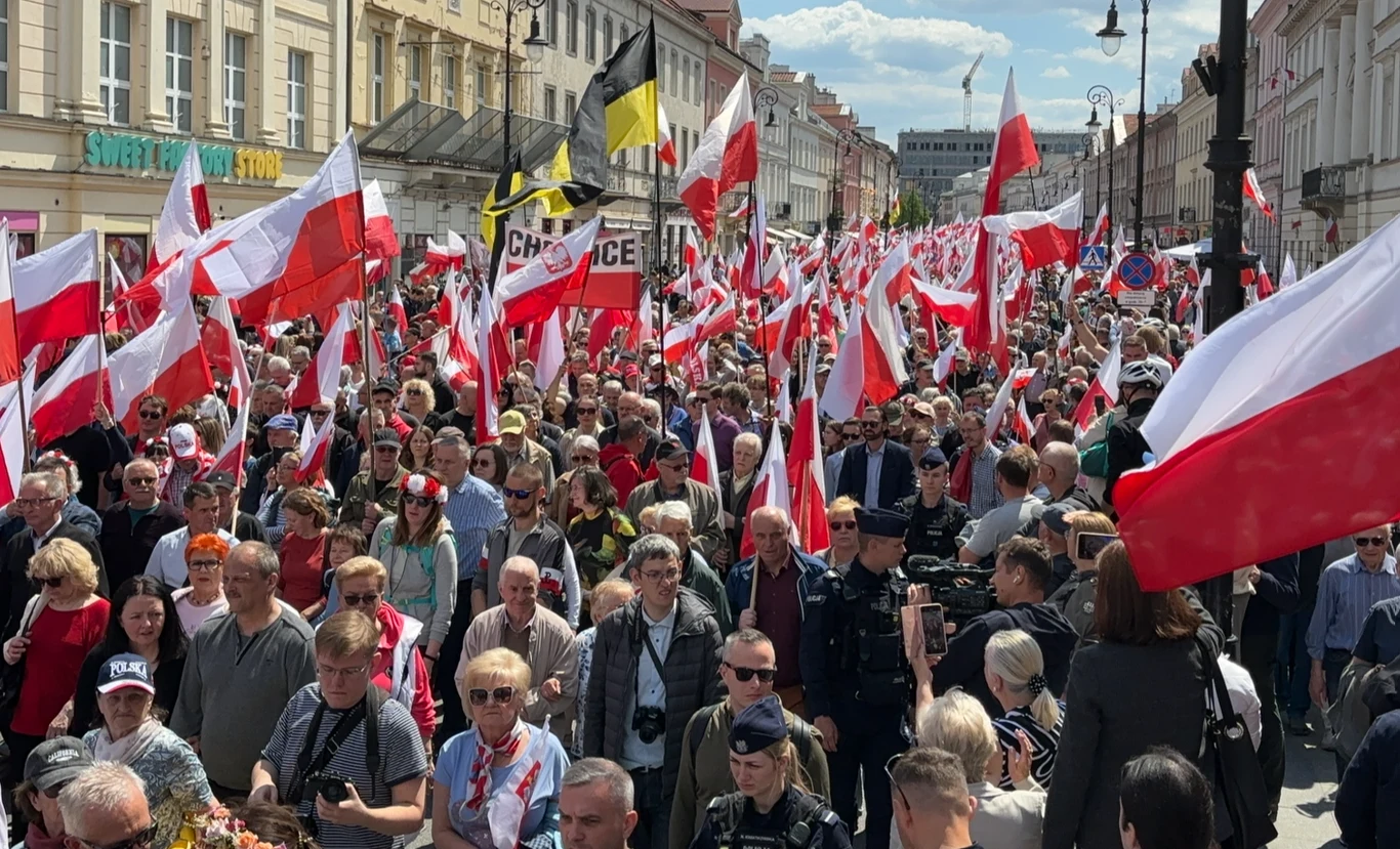 Tłum ludzi maszeruje ulicą, większość uczestników trzyma polskie flagi, nad głowami góruje morze biało-czerwonych barw, po bokach stoją policjanci, a w tle widoczne są kamienice i niebo z lekkim zachmurzeniem.