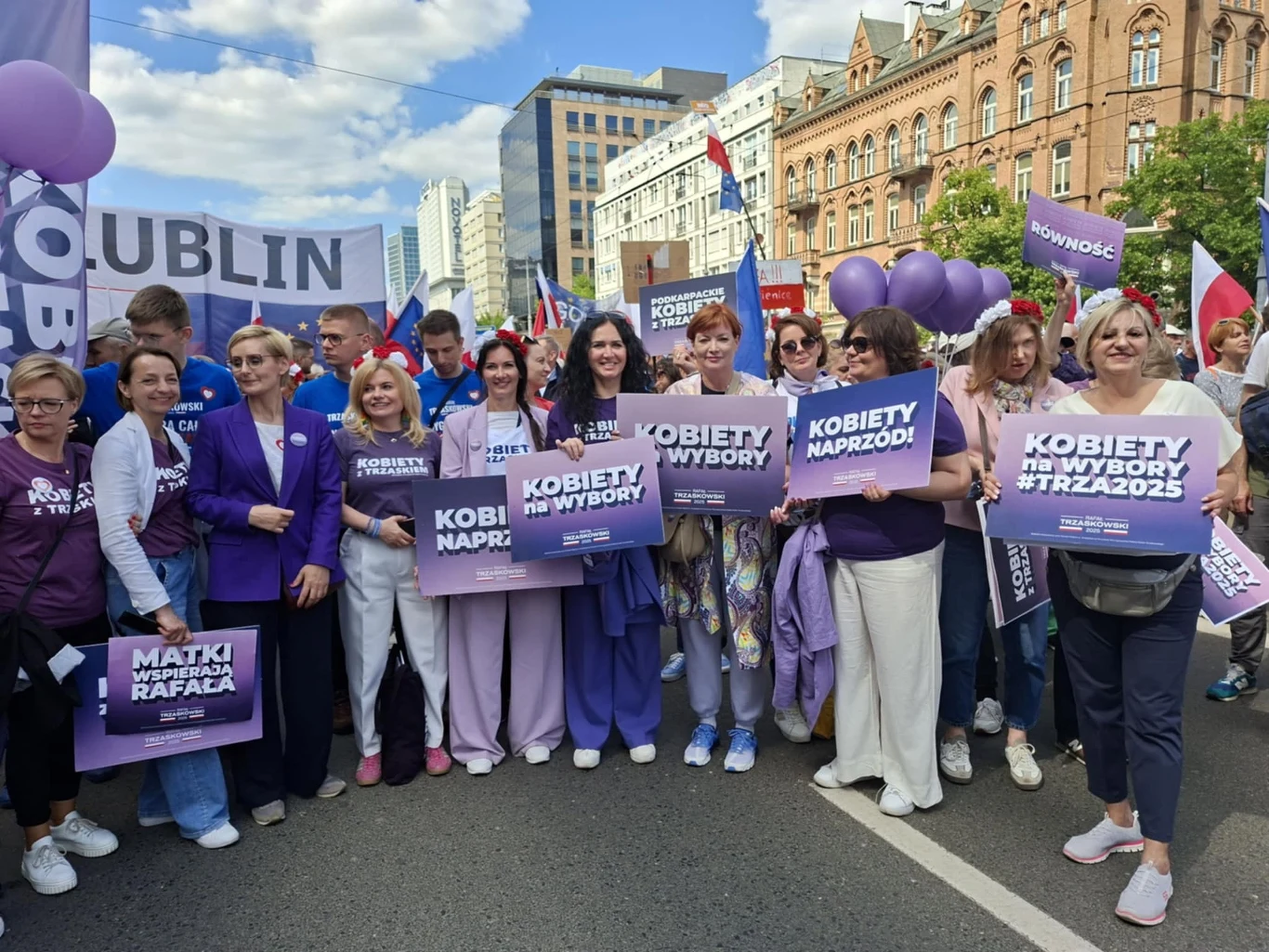 Grupa kobiet trzymających transparenty z hasłami dotyczącymi praw wyborczych kobiet, na tle miejskiej ulicy podczas marszu lub demonstracji w centrum miasta, otoczenie stanowią budynki oraz inni uczestnicy wydarzenia.