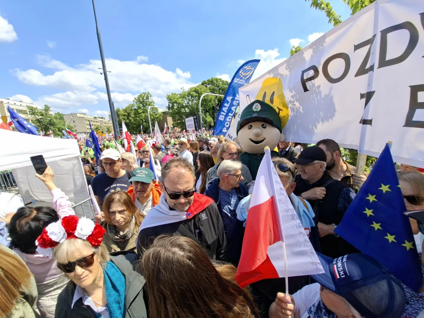 Tłum ludzi zgromadzonych podczas pokojowej manifestacji, wiele osób trzyma flagi Polski oraz Unii Europejskiej, widoczny jest także transparent z napisem oraz postać w dużym pluszowym przebraniu.
