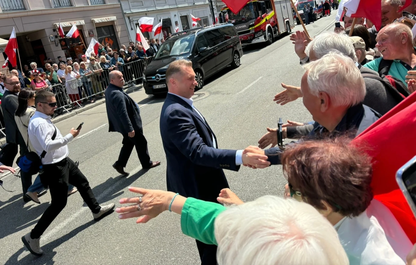 Polityk w garniturze ściska dłonie zgromadzonych ludzi podczas uroczystości ulicznej otoczonej przez tłum oraz flagi Polski, w tle widoczny samochód i inni uczestnicy wydarzenia.
