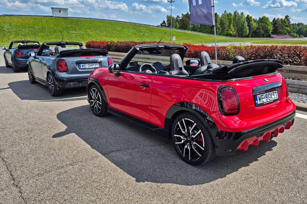 Mini JCW Cabrio to zdecydowanie najbardziej emocjonująca wersja w gamie Rząd kolorowych kabrioletów Mini Cooper ustawionych na asfalcie toru, w tle zielone trawniki i drzewa pod jasnym, letnim niebem.