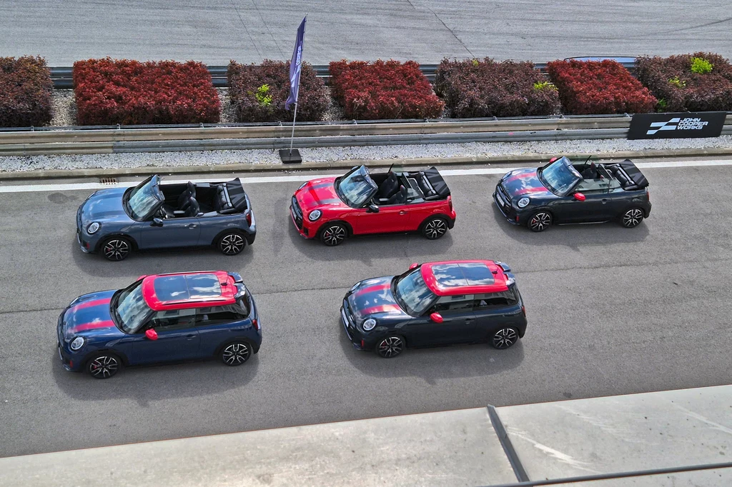 Mini John Cooper Works w wersji hatchback ma napęd spalinowy lub elektryczny, kabriolet tylko spalinowy Pięć kabrioletów Mini Cooper w nietypowych wersjach kolorystycznych zaparkowanych na torze wyścigowym, widok z góry, wokół znajduje się bariera oraz niskie krzewy.