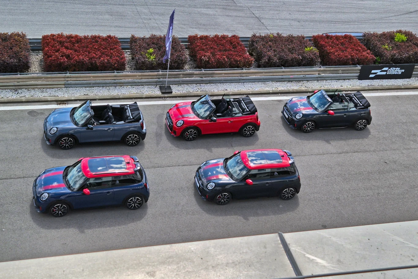 Pięć kabrioletów Mini Cooper w nietypowych wersjach kolorystycznych zaparkowanych na torze wyścigowym, widok z góry, wokół znajduje się bariera oraz niskie krzewy.