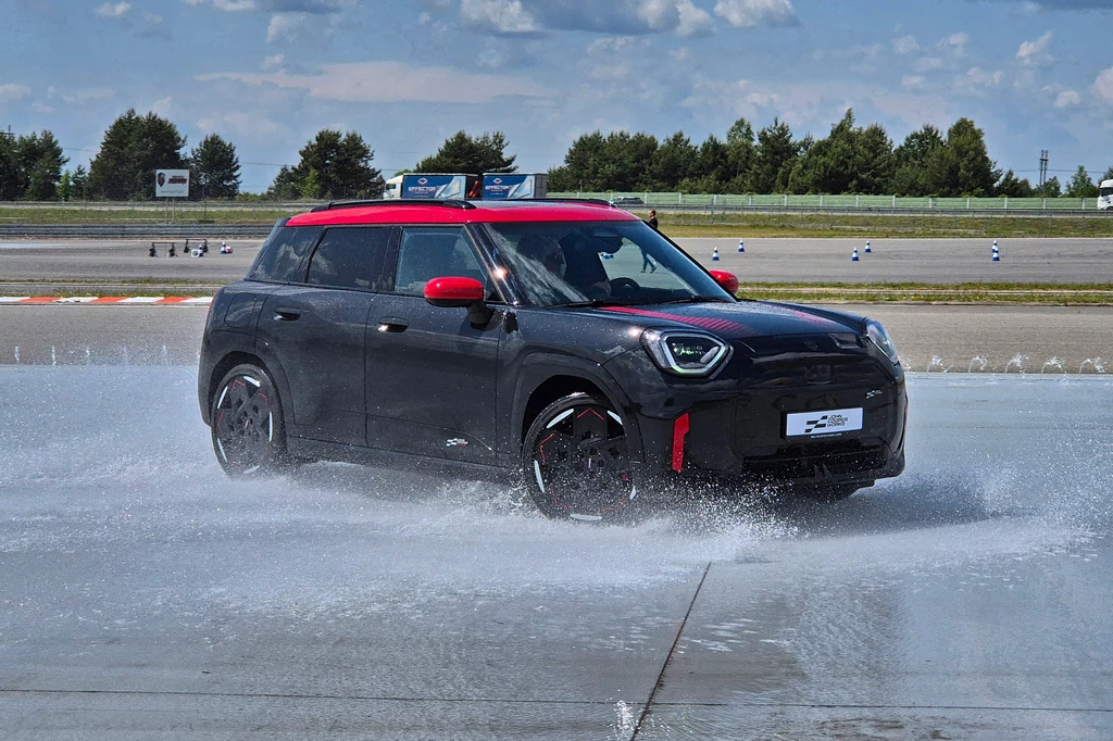 Mini JCW Aceman to model wyłącznie elektryczny Czarny samochód z czerwonym dachem i lusterkami jedzie po mokrej nawierzchni, rozpryskując wodę wokół siebie, w tle widoczne są drzewa i tor wyścigowy.