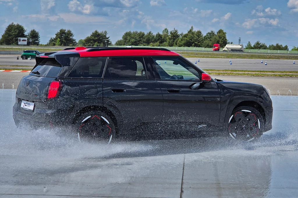 Mini JCW Aceman nie ma w sobie lekkości spalinowych Mini John Cooper Works Czarny samochód typu SUV z czerwonym dachem i lusterkami pokonuje zakręt na mokrej nawierzchni toru testowego, wokół pojazdu rozpryskuje się woda, w tle drzewa oraz fragmenty infrastruktury toru.