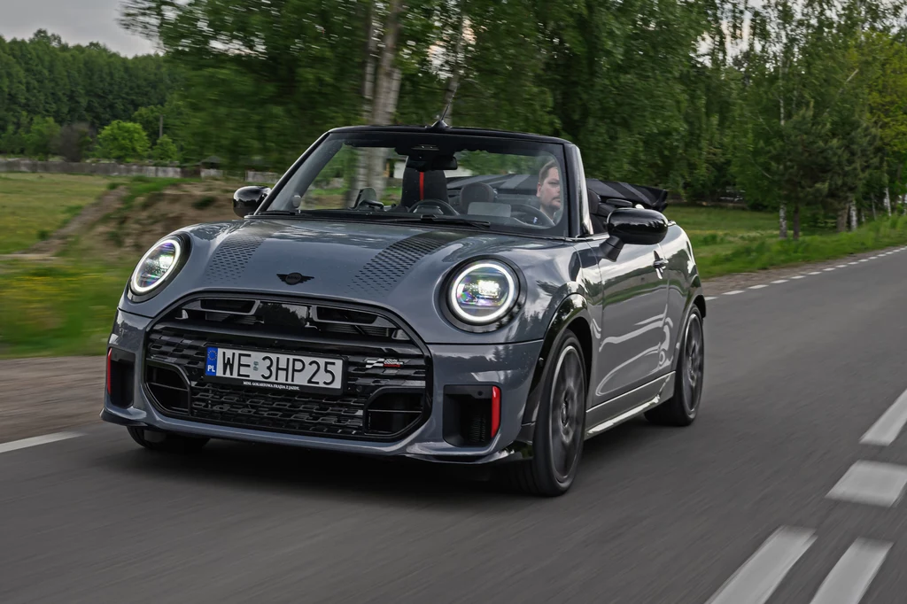 Mini JCW Cabrio to mój faworyt z całej gamy John Cooper Works Szary kabriolet marki Mini jedzie po asfaltowej drodze, otoczony zielenią i drzewami, dach samochodu jest otwarty, w środku znajduje się kierowca.