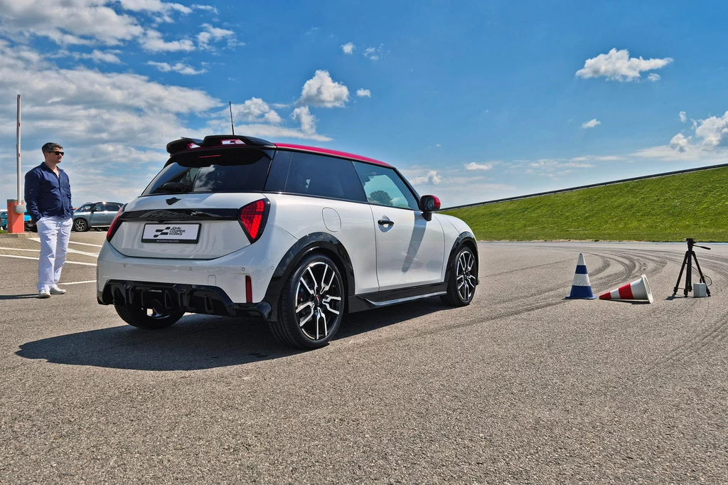 Charlie Cooper, wnuk Johna Coopera, brał udział w pierwszych testach nowych Mini JCW w Polsce Biały samochód typu hatchback z czerwonym dachem stoi na asfalcie przy torze testowym, w pobliżu rozstawione są kolorowe pachołki i statyw z urządzeniem pomiarowym, po lewej znajduje się mężczyzna w białych spodniach i ciemnej kurtce obserwujący sytuac...
