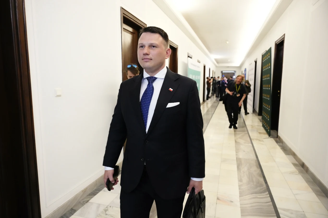 Piwo z Trzaskowskim. Sławomir Mentzen: Zaskoczyła mnie skala reakcji Elegancko ubrany mężczyzna w garniturze i krawacie z broszką w barwach narodowych przechodzi korytarzem urzędowym, trzymając w dłoni teczkę. W tle widoczni są inni ludzie poruszający się po tym samym korytarzu.