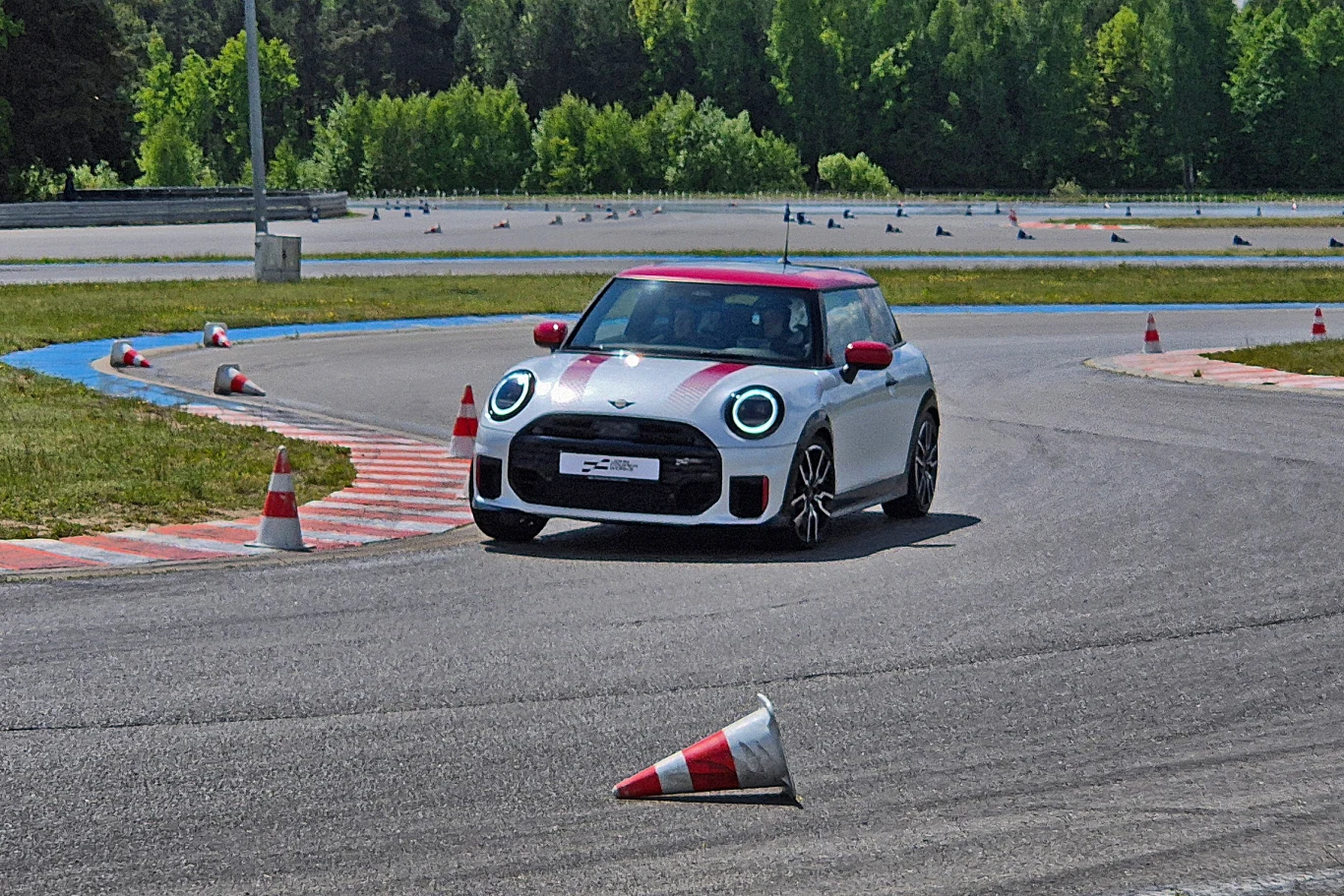 Samochód sportowy Mini Cooper w biało-czerwonych barwach pokonuje zakręt na torze wyścigowym, wokół rozstawione są pachołki zabezpieczające trasę, w tle bujna, zielona roślinność oraz asfaltowy tor.