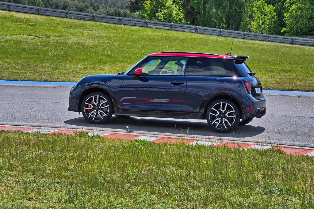 Mini John Cooper Works daje dużo frajdy na torze Mały sportowy samochód o granatowym lakierze z czerwonym dachem jedzie po torze wyścigowym, tło stanowi zielona trawa i niewielkie wzniesienia.