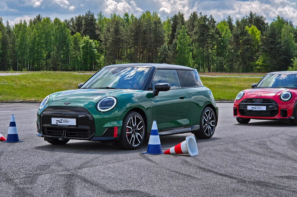 Mini JCW E, a za nim spalinowe Mini JCW Zielony samochód osobowy Mini Cooper ustawiony na asfalcie pomiędzy niebiesko-białymi i pomarańczowo-białymi pachołkami, w tle widoczny drugi czerwony samochód tego samego modelu, otoczenie stanowi zielony teren oraz ściana lasu pod lekko zachmurzonym ...