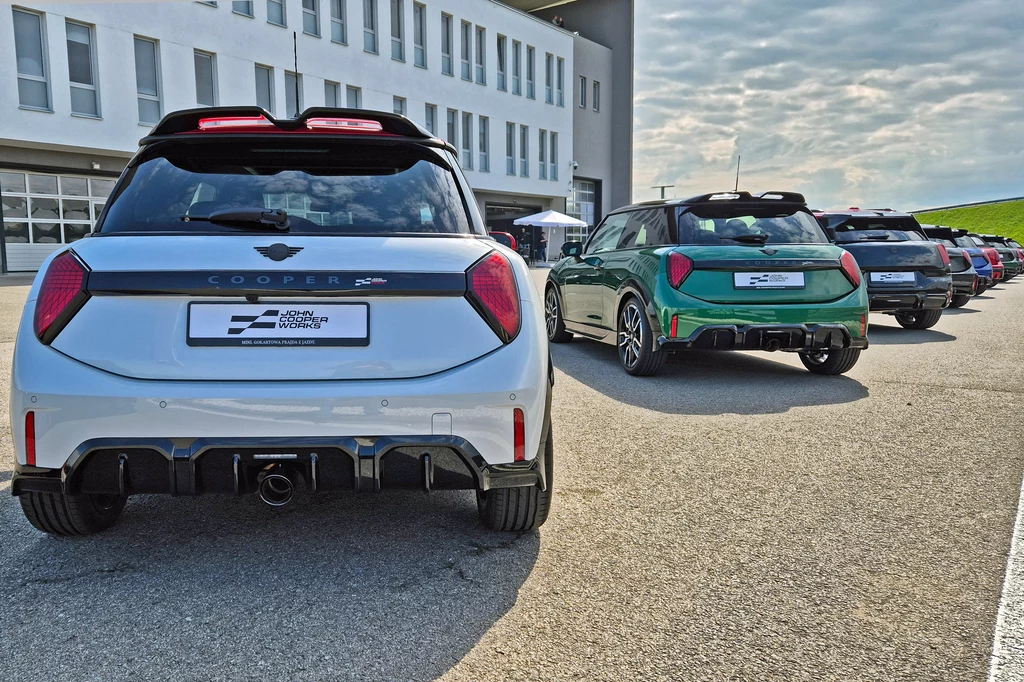 Mini JCW dostępne są w wersjach spalinowej i elektrycznej Nowoczesne, sportowe samochody MINI Cooper ustawione w szeregu na asfaltowym placu przy nowoczesnym budynku, wyróżniają się aerodynamiczną sylwetką i charakterystycznymi tylnymi światłami; z tyłu widoczny napis Cooper oraz logo MINI.