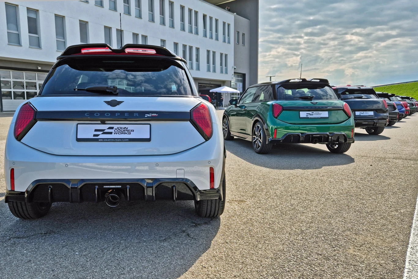 Nowoczesne, sportowe samochody MINI Cooper ustawione w szeregu na asfaltowym placu przy nowoczesnym budynku, wyróżniają się aerodynamiczną sylwetką i charakterystycznymi tylnymi światłami; z tyłu widoczny napis Cooper oraz logo MINI.