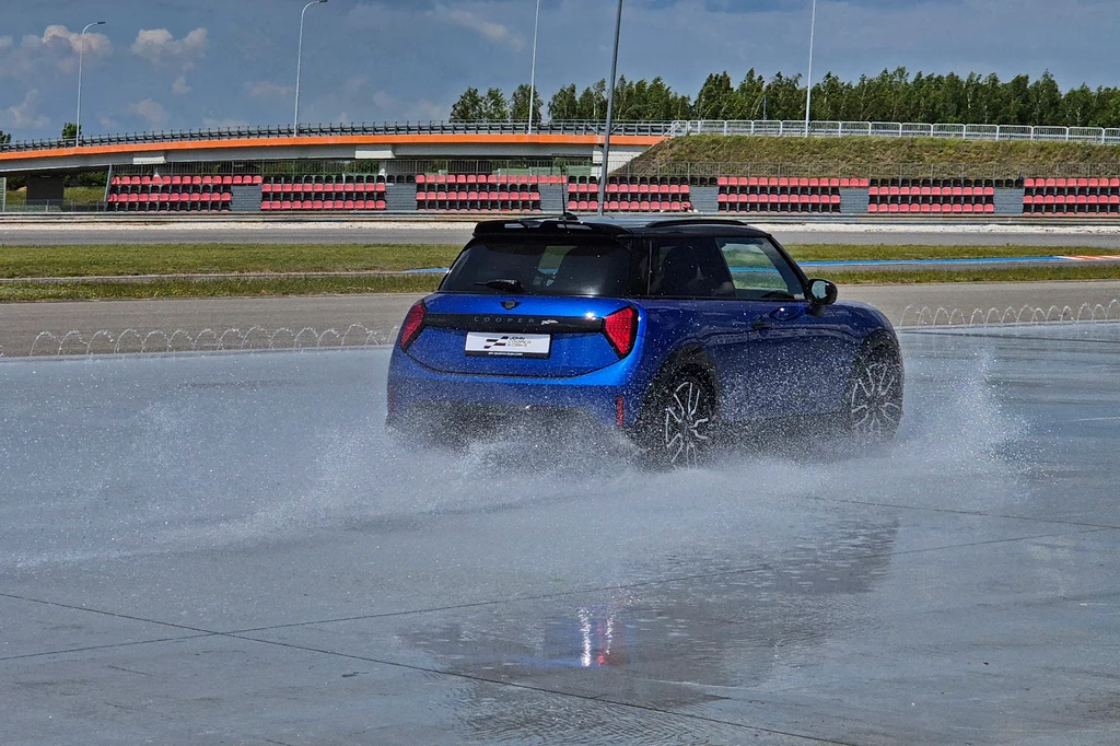 Spalinowe Mini JCW pozwala się bardzo łatwo kontrolować, również po wpadnięciu w poślizg Niebieski samochód osobowy wykonuje test poślizgu na mokrej nawierzchni na specjalnym torze, wokół widoczne są rozpryski wody oraz infrastruktura toru testowego z trybunami w tle.