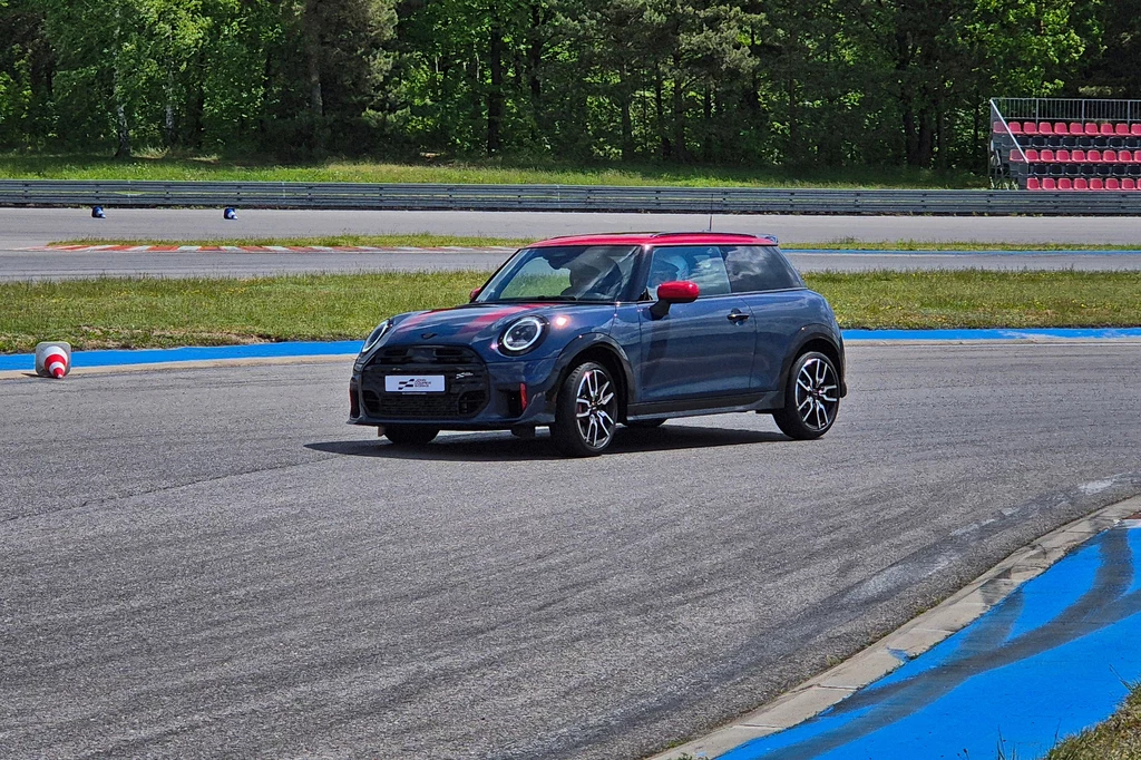 Mini JCW Samochód Mini Cooper w kolorze czarnym z czerwonym dachem pokonuje zakręt na torze wyścigowym, wokół widoczne są barierki ochronne i zielone drzewa w tle.