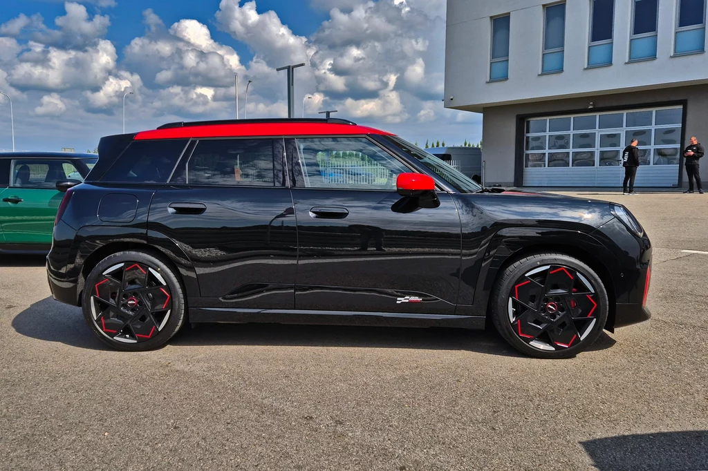 Mini JCW Aceman Czarny samochód typu SUV z czerwonymi detalami zaparkowany na asfaltowym placu, w tle budynek o nowoczesnej architekturze oraz kilka osób stojących przy ścianie.