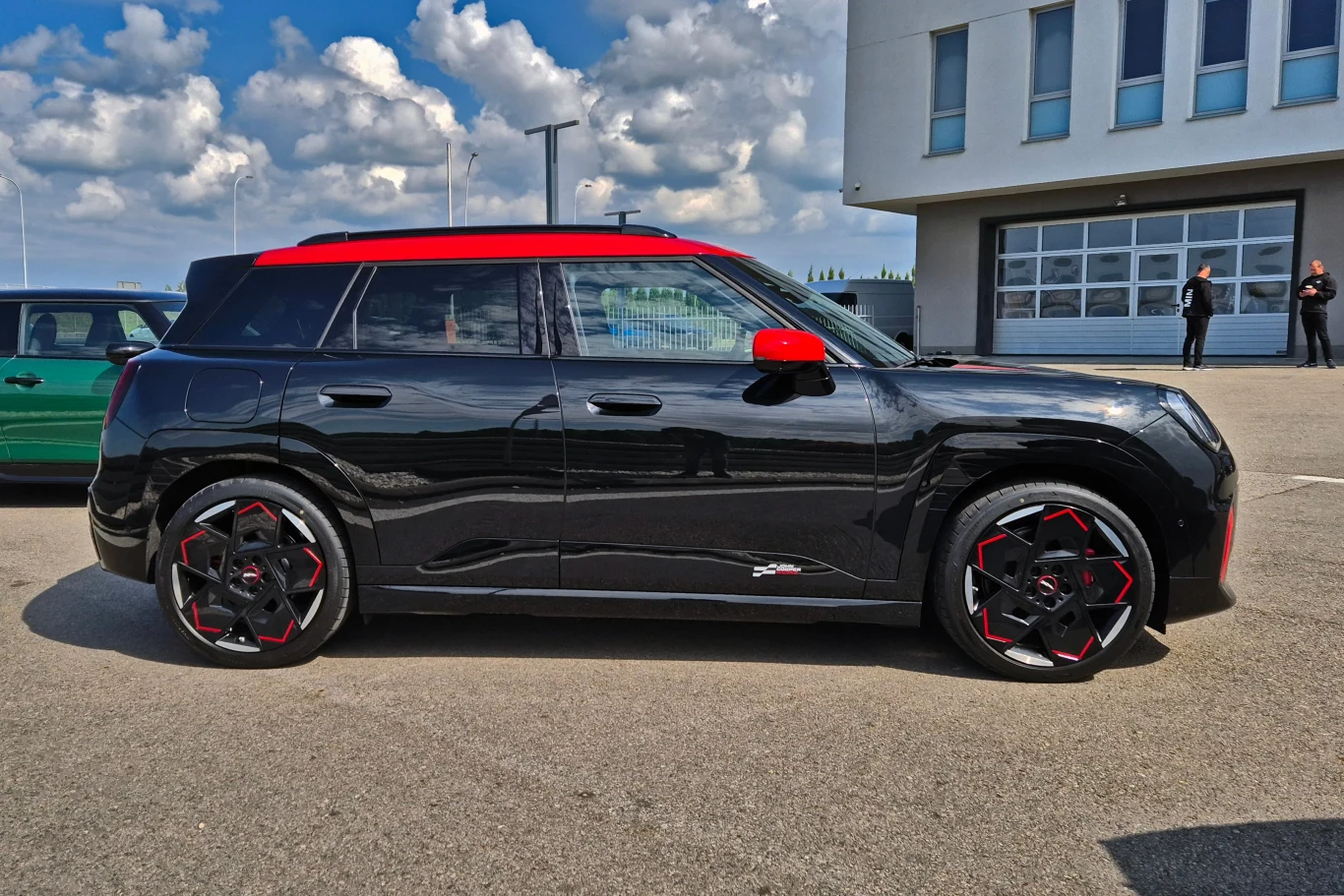 Czarny samochód typu SUV z czerwonymi detalami zaparkowany na asfaltowym placu, w tle budynek o nowoczesnej architekturze oraz kilka osób stojących przy ścianie.