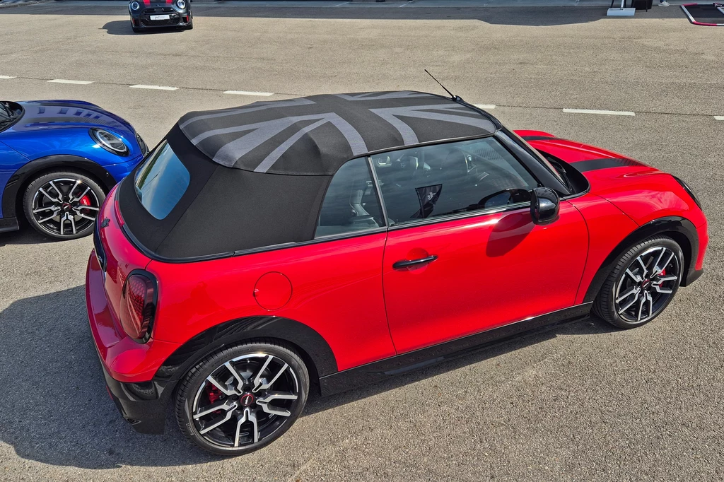 Mini JCW Cabrio Czerwony kabriolet Mini Cooper z miękkim dachem z motywem brytyjskiej flagi, zaparkowany na asfaltowej nawierzchni obok niebieskiego samochodu tego samego modelu.