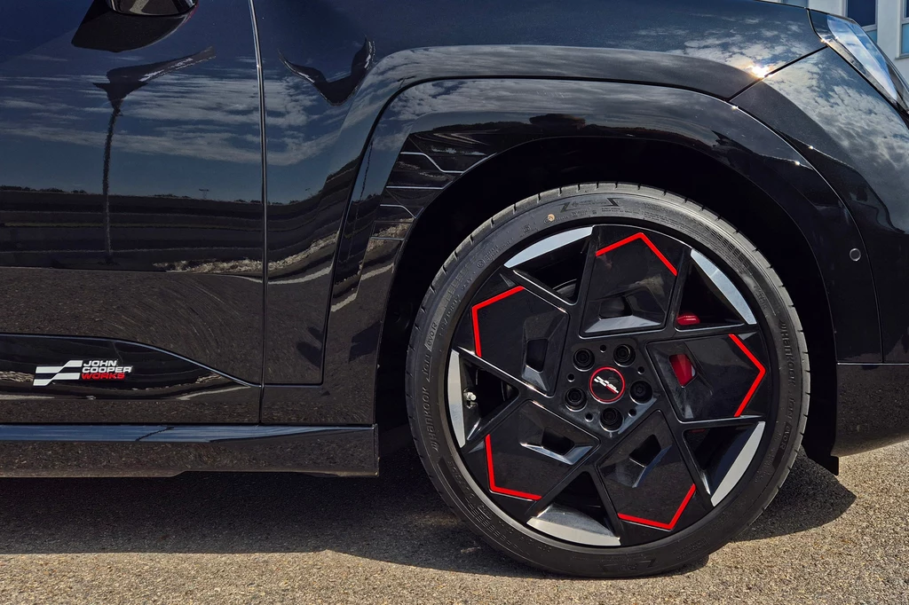 Mini JCW Aceman Czarna karoseria samochodu sportowego z widocznym nowoczesnym kołem aluminiowym ozdobionym czerwonymi akcentami, asfaltowa nawierzchnia drogi i zarys linii na karoserii.