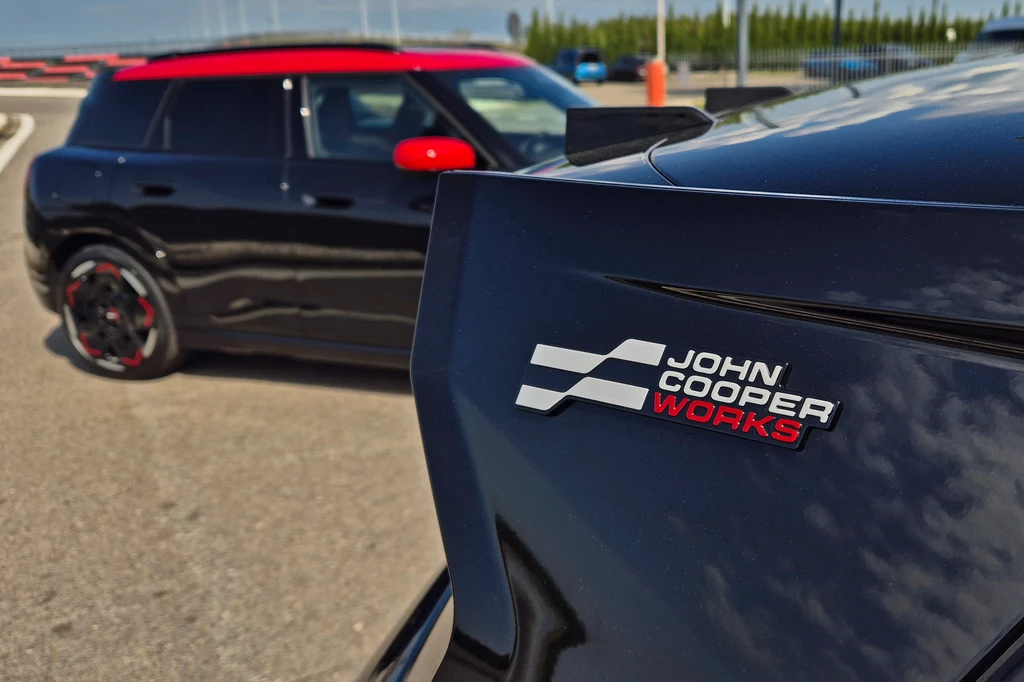 Mini JCW E Czarny samochód Mini John Cooper Works z czerwonym dachem i lusterkami stoi na torze wyścigowym, na pierwszym planie widoczny znaczek modelu na tylnej części innego auta.