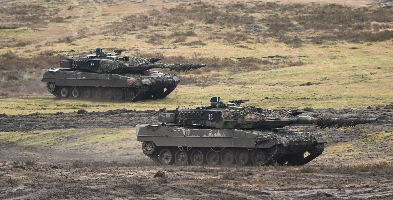Niemieckie czołgi Leopard 2. Zdjęcie ilustracyjne Dwa niemieckie czołgi Leopard 2 przemieszczające się po otwartym, błotnistym terenie, pomalowane w kamuflaż leśny, w tle sucha, trawiasta przestrzeń.