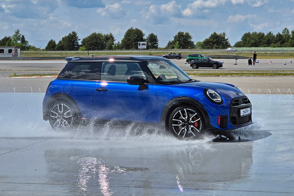 Mini JCW Niebieski samochód Mini Cooper jedzie po mokrej nawierzchni, rozpylając wodę wokół kół, w tle asfaltowy tor oraz kilka innych pojazdów zaparkowanych lub jadących.