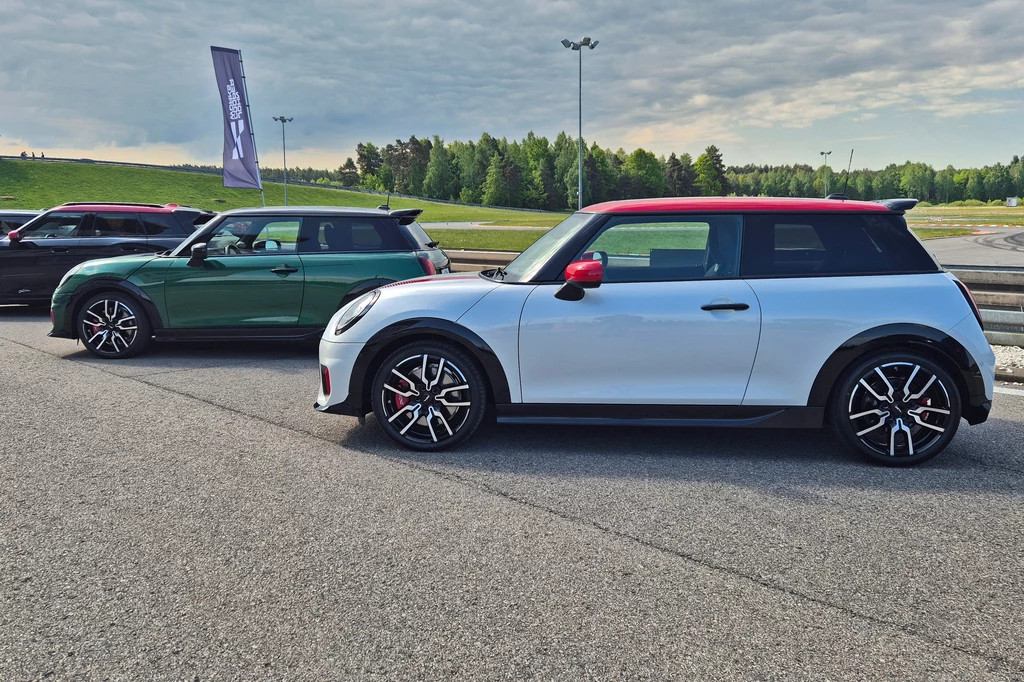 Mini JCW Dwa samochody Mini John Cooper Works zaparkowane obok siebie na asfalcie, widoczne na tle trawiastych pagórków i zielonych drzew, jeden samochód biały z czerwonym dachem, drugi zielony