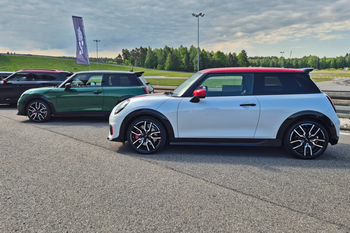 Dwa samochody Mini John Cooper Works zaparkowane obok siebie na asfalcie, widoczne na tle trawiastych pagórków i zielonych drzew, jeden samochód biały z czerwonym dachem, drugi zielony