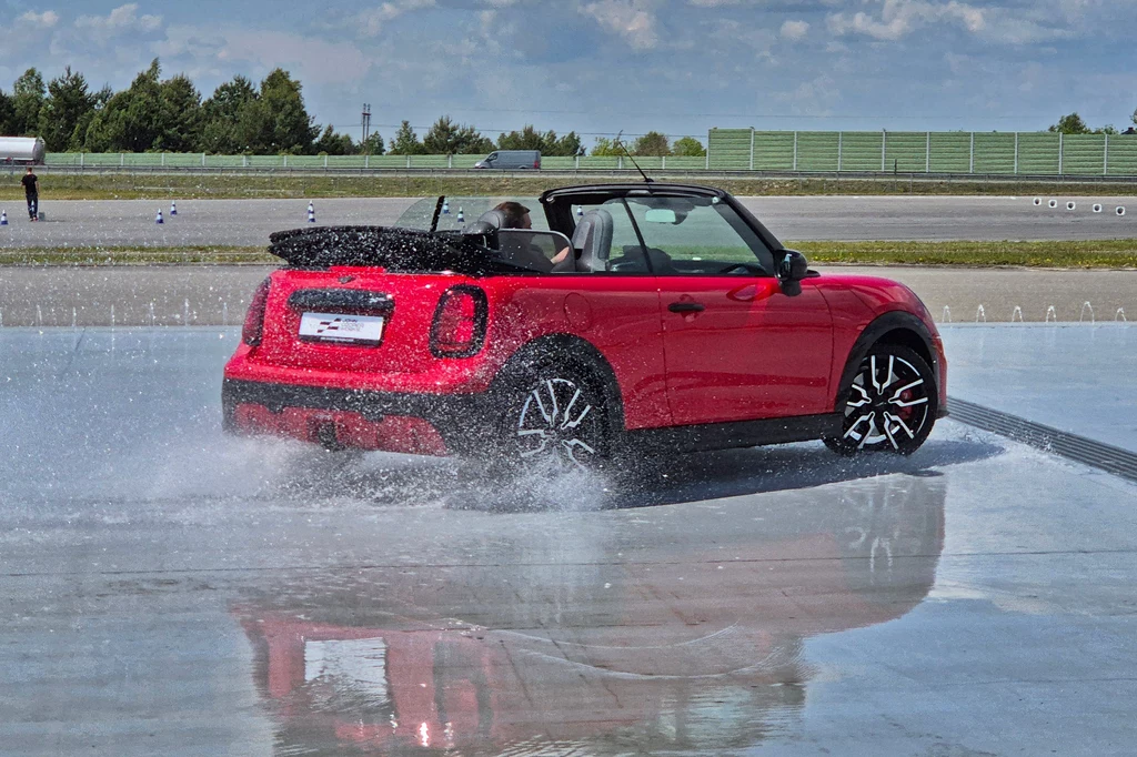 Mini JCW Cabrio Czerwony kabriolet podczas dynamicznej jazdy po torze z mokrą nawierzchnią, obfity rozprysk wody spod kół, samochód podczas testowania przyczepności lub symulacji poślizgu, otwarte dach i wyraźny ruch w lewo.