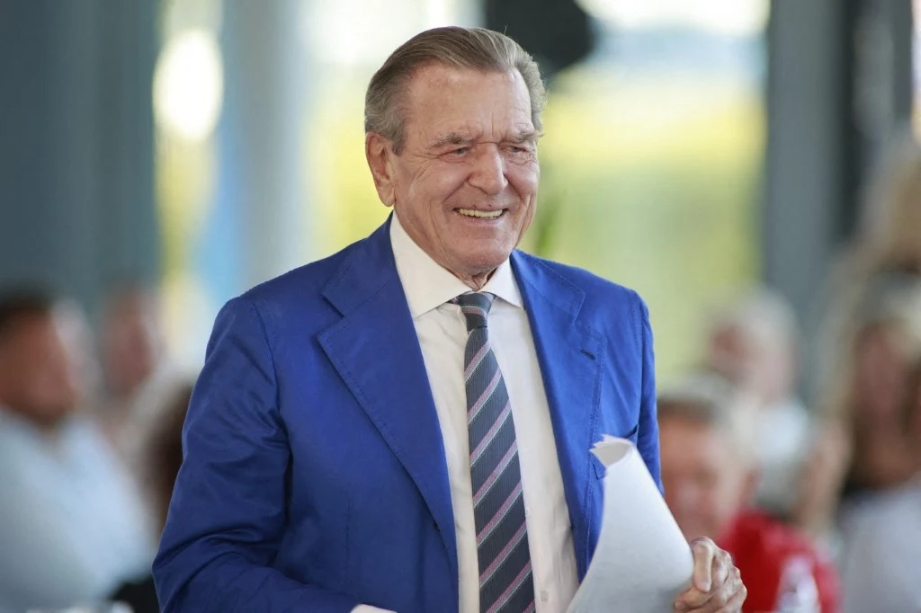 Bank zablokował konto Gerharda Schroedera Starszy mężczyzna w niebieskiej marynarce i krawacie w paski trzymający kartkę papieru, uśmiechnięty, w tle rozmyta grupa ludzi.
