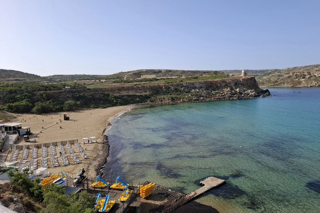 Plaża Golden Bay Szeroka, piaszczysta plaża z rzędem leżaków oraz parasoli po lewej stronie, molo i dmuchane łódki przy brzegu, błękitna, krystaliczna woda i strome, skaliste wzgórze w tle, częściowo pokryte zielenią.