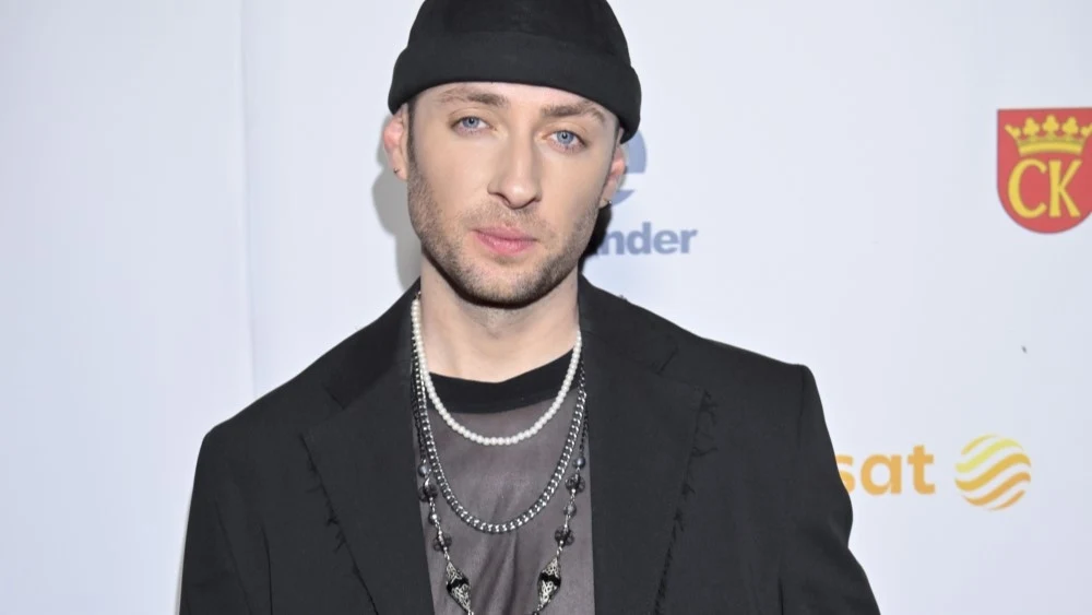 Dominik Dudek Mężczyzna w czarnej czapce, czarnej marynarce i przezroczystej koszulce, ozdobiony kilkoma naszyjnikami, stoi na tle ścianki sponsorskiej z widocznymi logotypami i herbem CK.