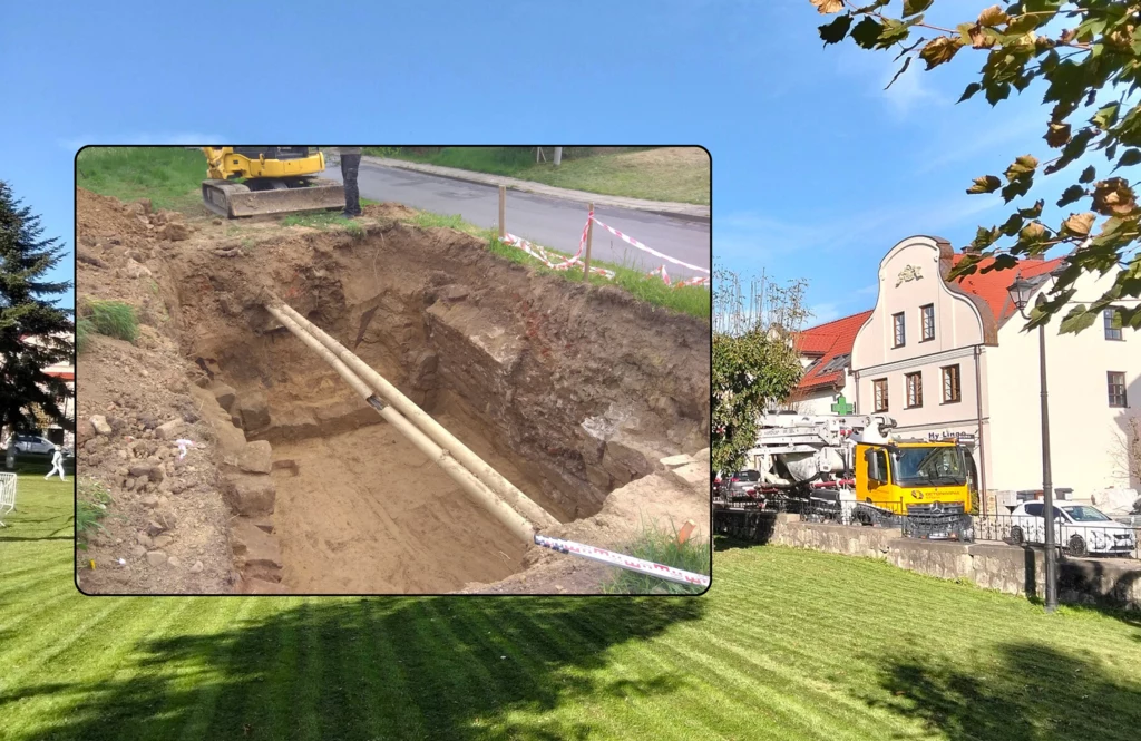 Na terenie Nowego Wiśnicza dokonano ciekawego odkrycia. Depozyt był ukryty głęboko pod ziemią. Wykop z rurami wodociągowymi podczas prac ziemnych, w tle widoczny koparka i osoba nadzorująca, całość zmontowana na tle miejskiego placu z wypielęgnowanym trawnikiem i budynkiem z białą fasadą.