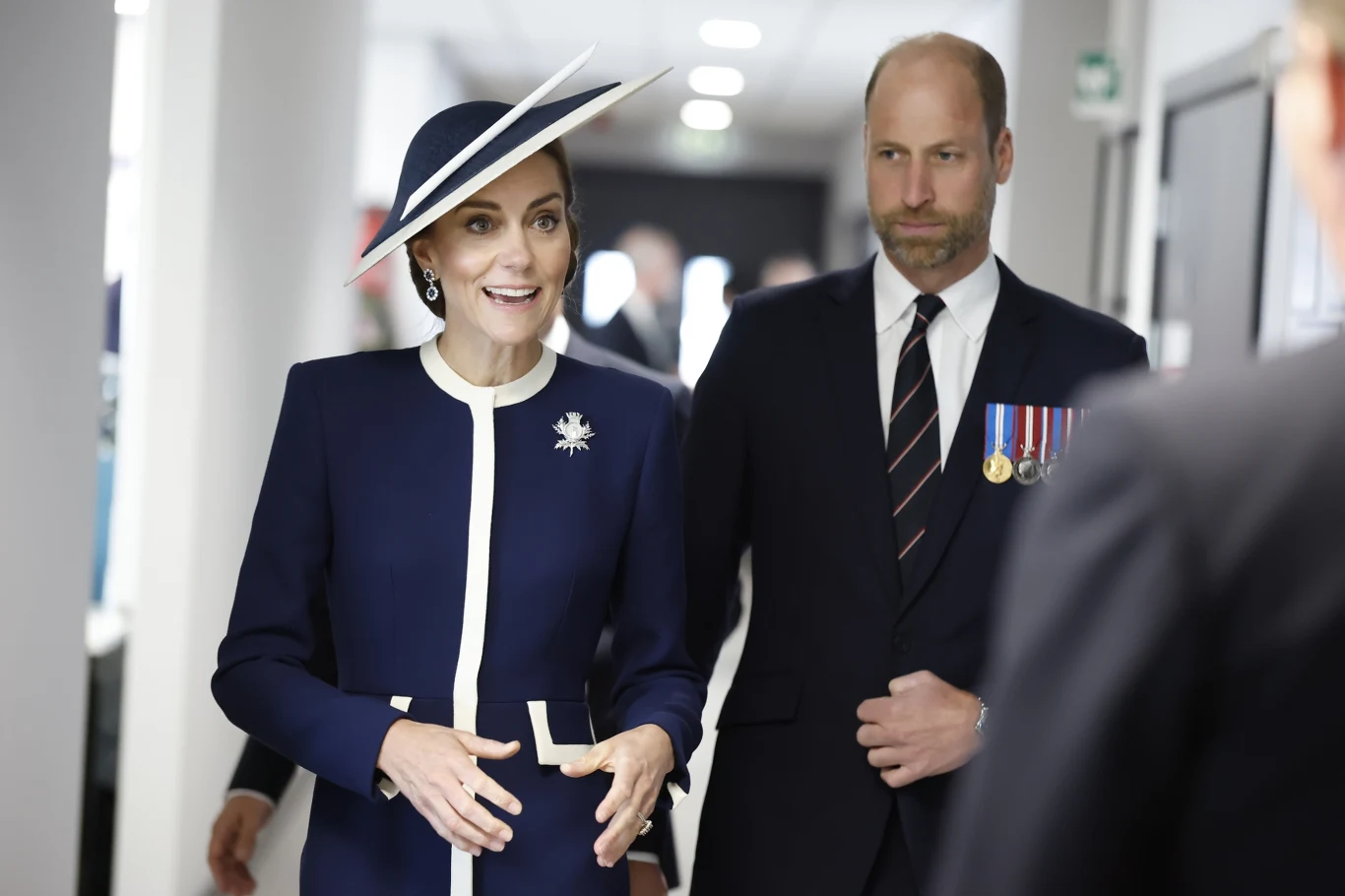 Księżna Kate zdecydowanie skupiła na sobie uwagę Kobieta w eleganckim granatowym kostiumie z białymi akcentami i szerokim kapeluszem stoi obok mężczyzny w garniturze z przypiętymi medalami, obydwie osoby uśmiechnięte, w tle jasny korytarz.