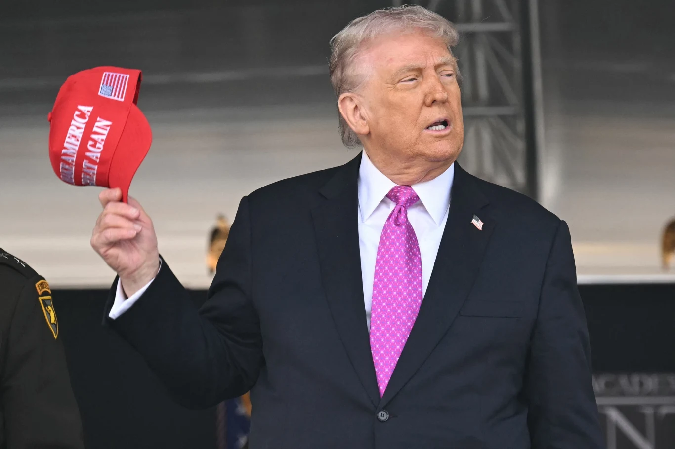 Przywracanie wielkości Ameryce na razie Trumpowi nie idzie tak, jak zaplanował Mężczyzna w garniturze i różowym krawacie trzyma w ręku czerwoną czapkę z napisem, w tle widoczne są fragmenty uniformów wojskowych oraz metalowa konstrukcja.