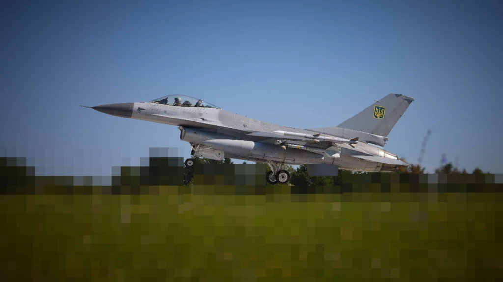 Samolot myśliwski F-16 w barwach ukraińskich podczas startu lub lądowania na pasie lotniska, podwozie opuszczone, tło stanowi pas zieleni i rozmyte niebo.