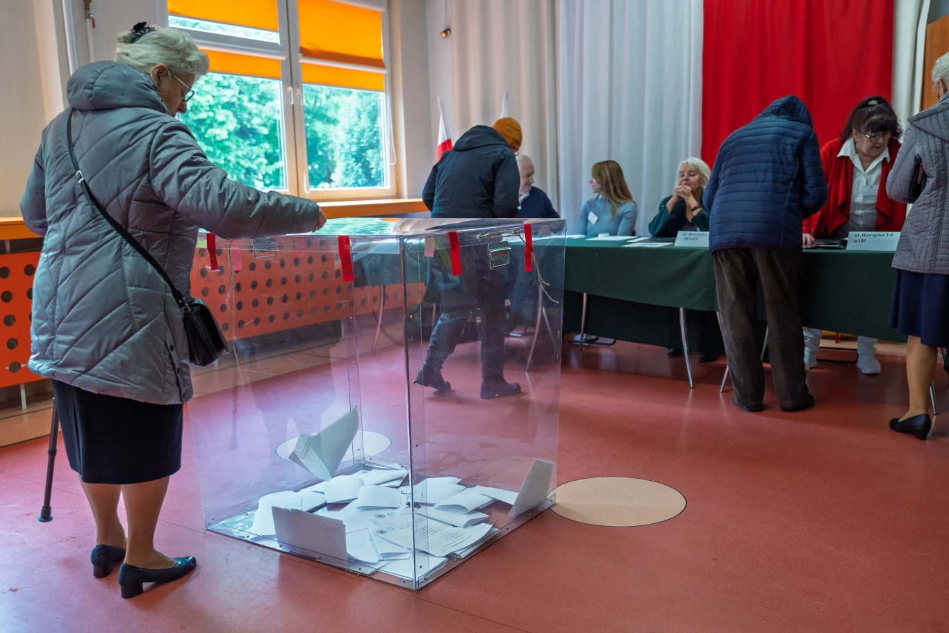 Wybory prezydenckie 2025, głosowanie Osoby oddające głosy przy przezroczystej urnie wyborczej w pomieszczeniu komisji wyborczej, przy stoliku siedzi kilku członków komisji, wnętrze oświetlone dziennym światłem, w tle czerwona zasłona.