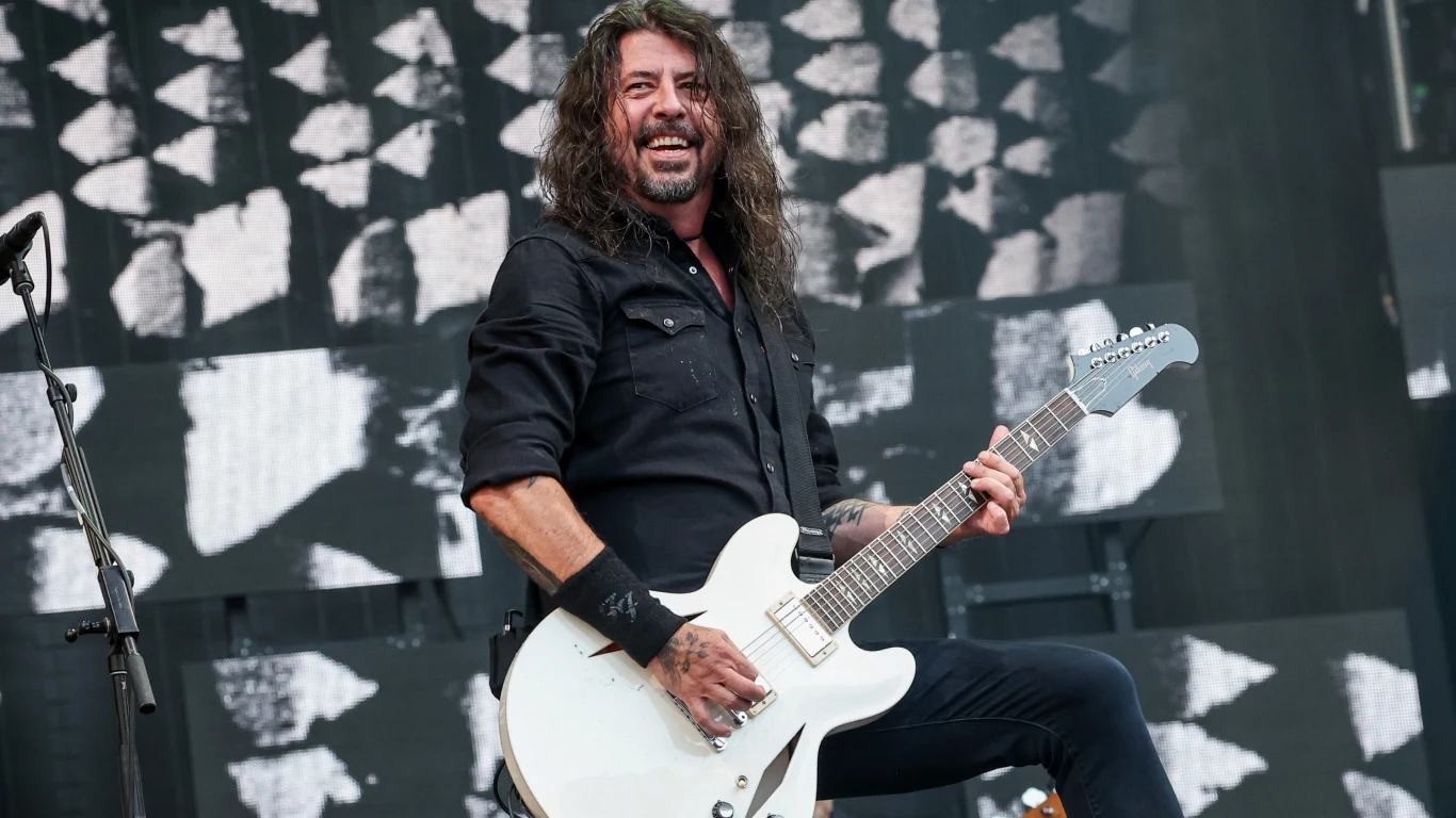 Foo FIghters Mężczyzna w czarnej koszuli z długimi włosami grający na białej gitarze elektrycznej na tle sceny koncertowej z abstrakcyjną czarno-białą grafiką.