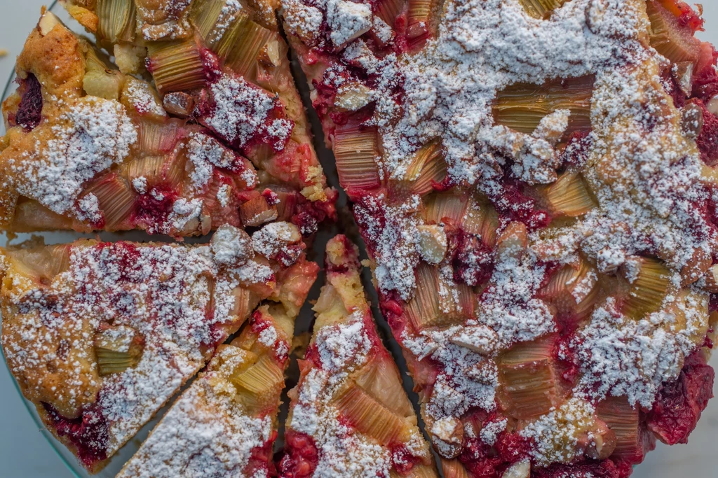 Ciasto z rabarbarem Kawałki ciasta z rabarbarem i owocami, obficie posypane cukrem pudrem, pokrojone na kilka porcji, widoczne kawałki rabarbaru i soczystych, czerwonych owoców.