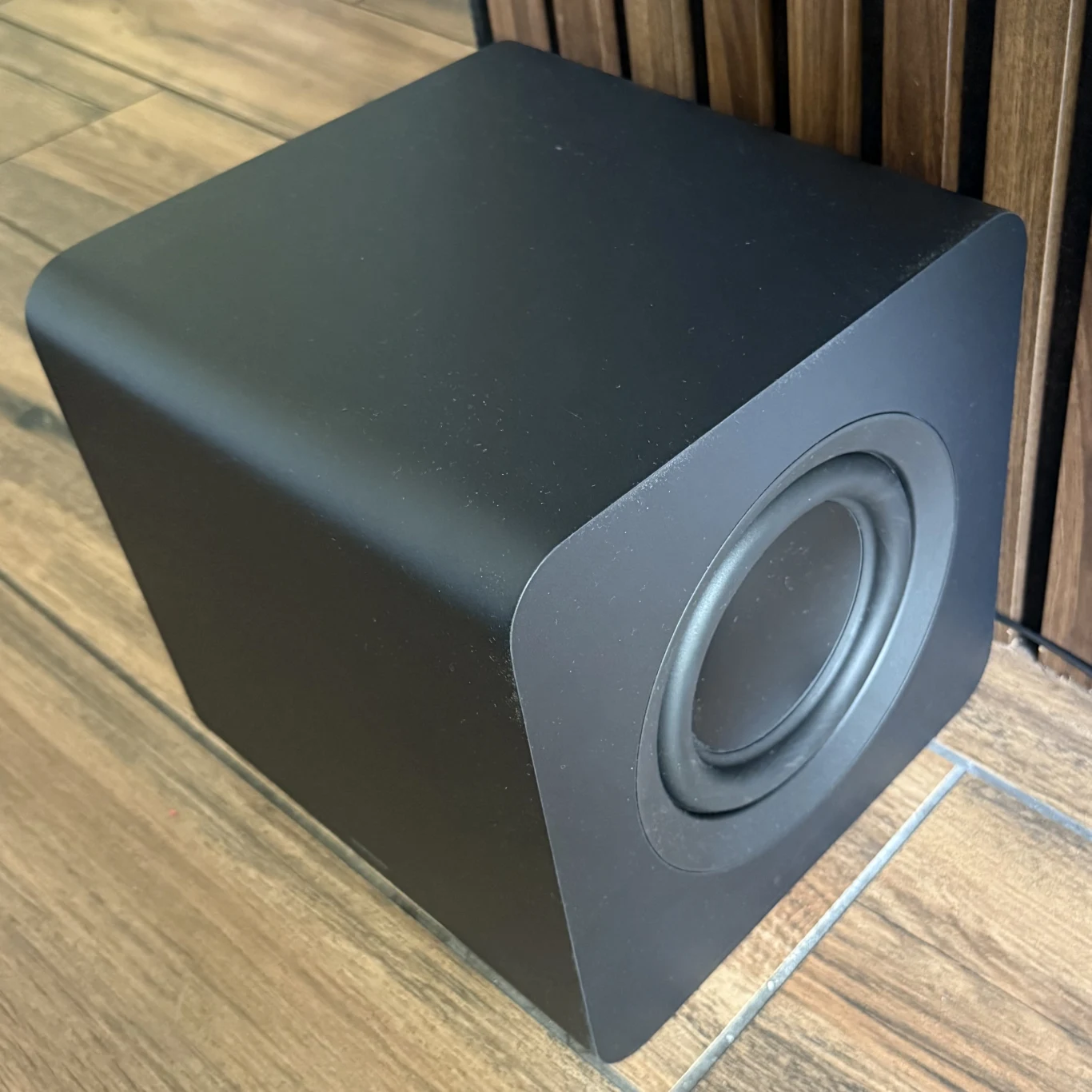 Subwoofer znajdujący się w zestawie z HW-Q930F jest znacznie mniejszy względem poprzednika. Czarny subwoofer o nowoczesnym, prostokątnym kształcie z zaokrąglonymi krawędziami ustawiony na drewnianej podłodze obok paneli ściennych.