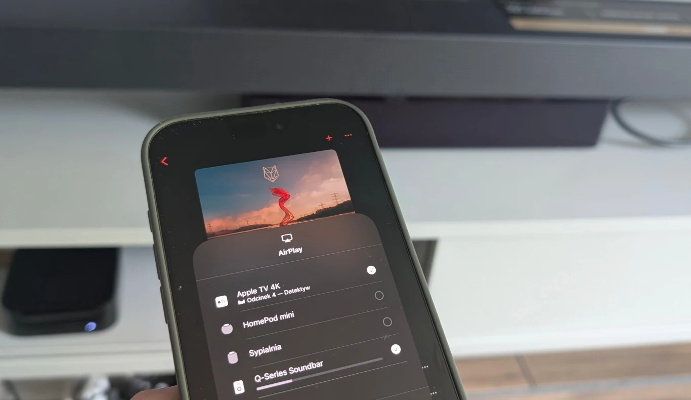 Użytkownicy sprzętów Apple z pewnością docenią wsparcie dla AirPlay. Ekran smartfona prezentujący menu AirPlay umożliwiający wybór urządzenia do odtwarzania multimediów, na tle widoczny fragment telewizora i półki.