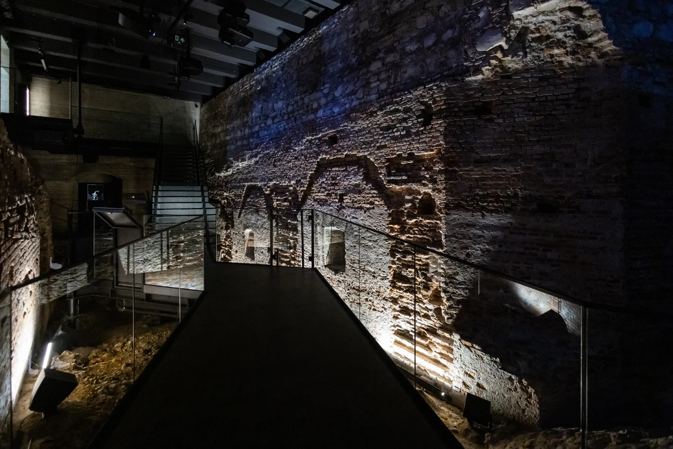 Oświetlony fragment starożytnego muru z widocznymi szczegółami ceglanej konstrukcji, metalowa kładka prowadząca przez historyczne miejsce, subtelne światła podkreślające fakturę ściany.