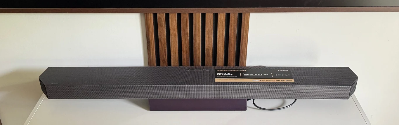 Soundbar HW-Q930F najwięcej możliwości oferuje w połączeniu z nowymi telewizorami marki Samsung, Soundbar marki Samsung ustawiony na białej szafce, tuż pod drewnianą listwą dekoracyjną, z widocznym przewodem zasilającym.