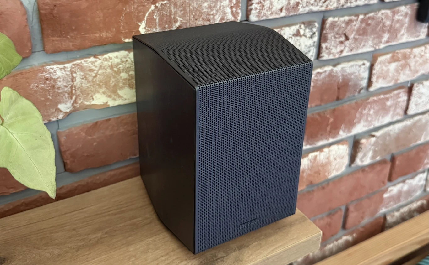 W zestawie z soundbarem Samsung HW-Q930F znajdują się także dwa dodatkowe głośniki. Czarny, nowoczesny głośnik bezprzewodowy ustawiony na drewnianej półce przy ścianie z czerwonej cegły, po lewej stronie widoczny fragment zielonej rośliny.