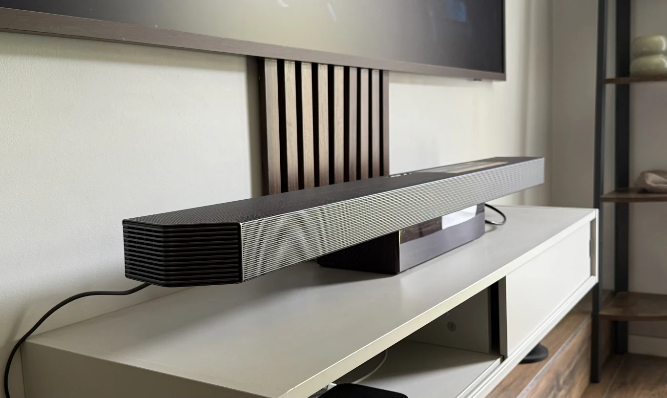 Soundbar Samsung HW-Q930F. Soundbar ustawiony na białej szafce RTV pod ściennym telewizorem, na tle dekoracyjnych, pionowych listew przytwierdzonych do ściany, z widoczną częścią pokoju i półką po prawej stronie.
