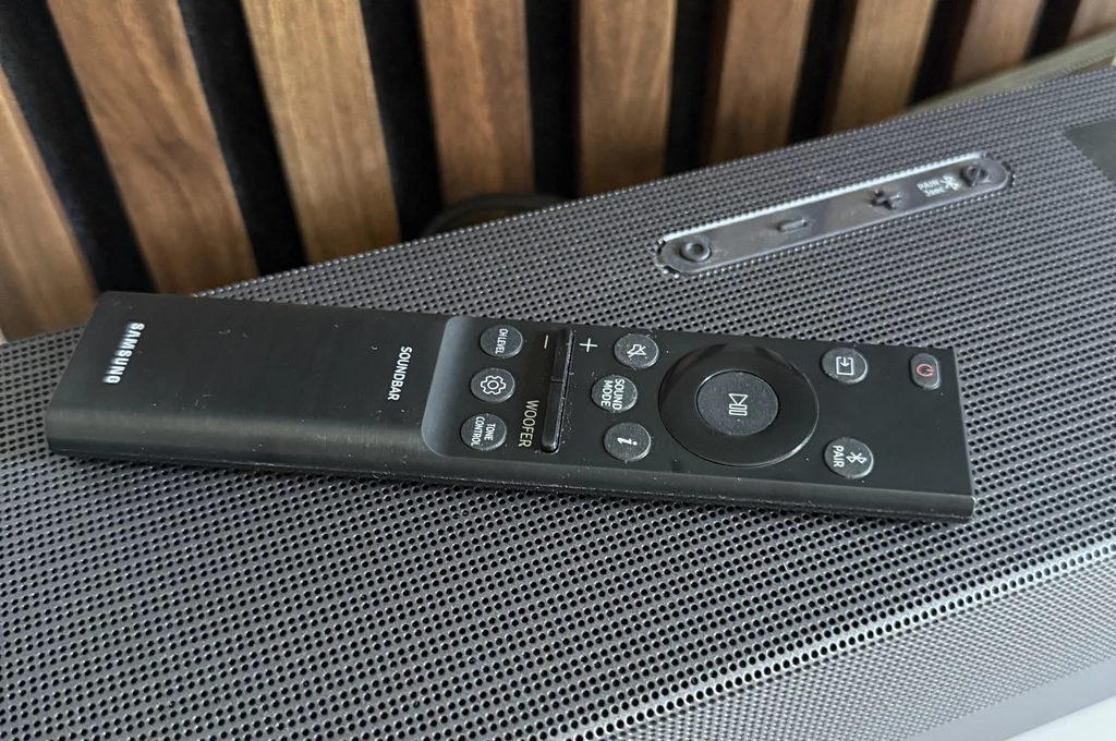 W zestawie jest pilot zdalnego sterowania, który pozwala także na kontrolowanie telewizora. Czarny pilot do sprzętu audio Samsung leży na kratowanej powierzchni soundbaru, w tle widoczne są drewniane pionowe panele.